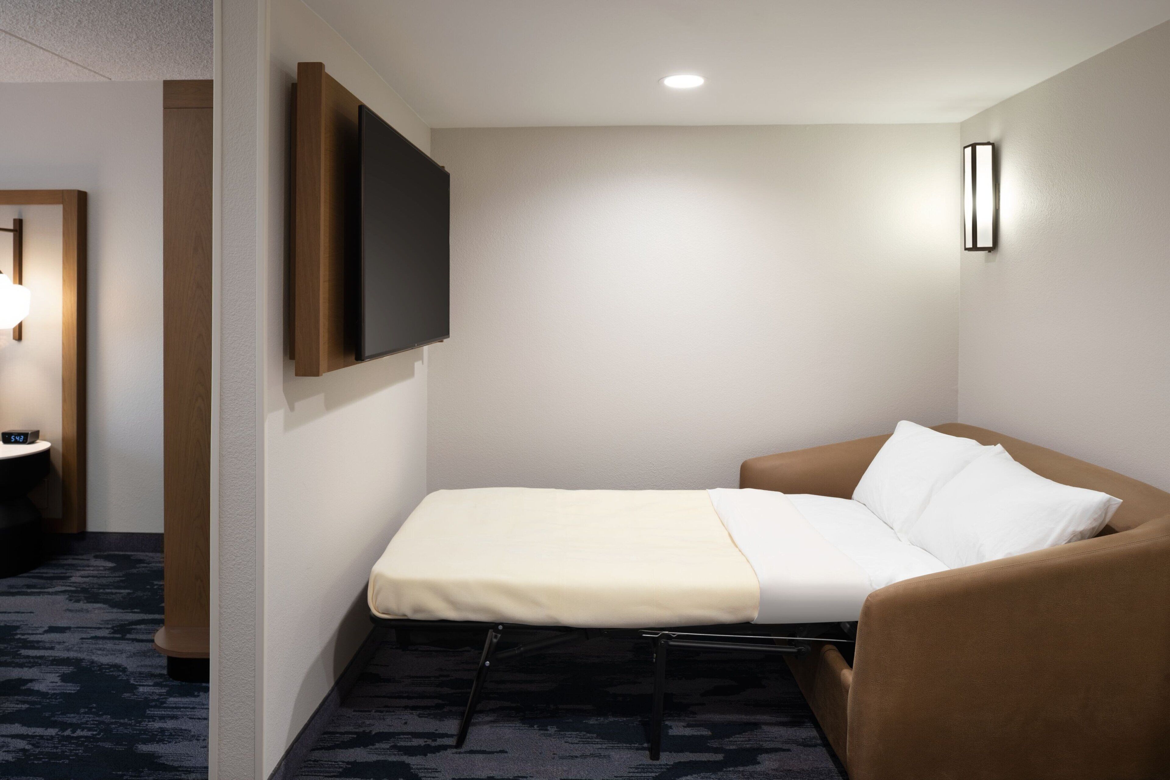 Een hotelkamer met een enkel bed, een flatscreen televisie aan de muur en een nachtkastje met een lamp.