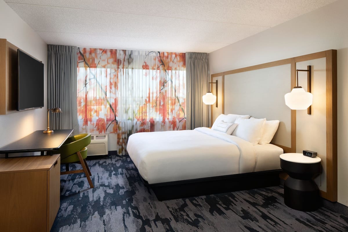 A hotel room with a bed, a desk, a chair, a TV, and a bedside table with a clock.