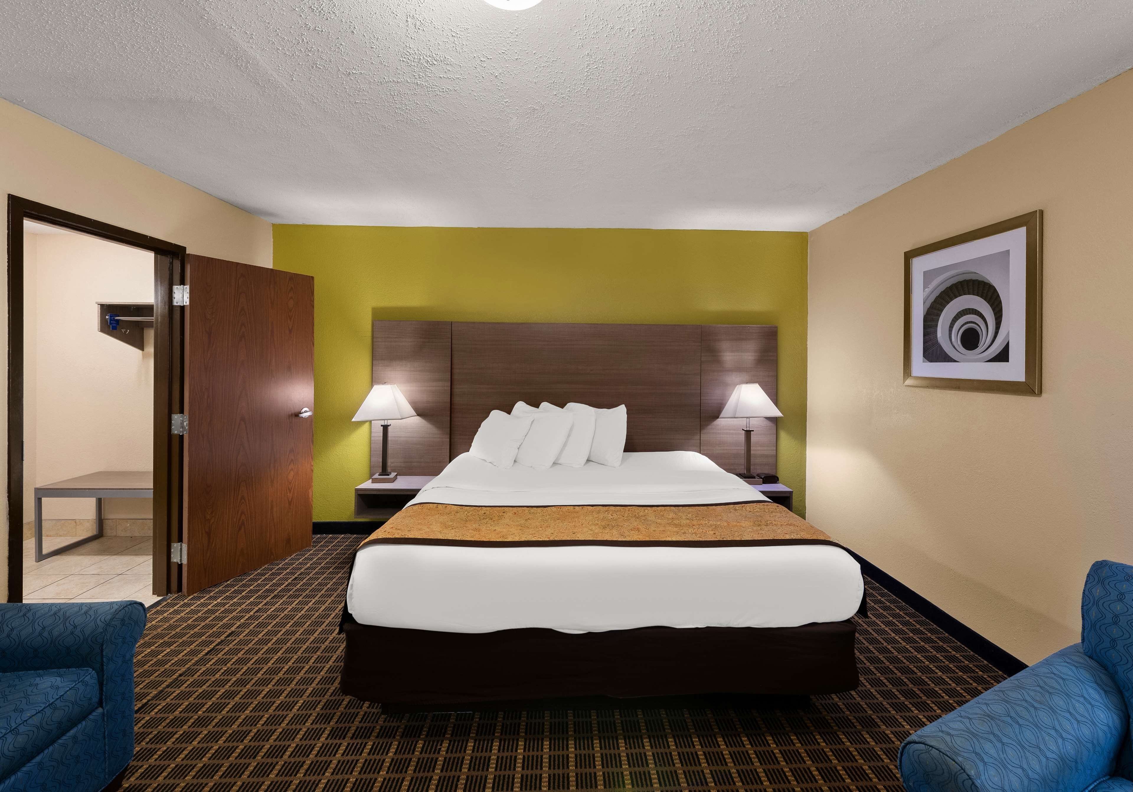 A hotel room with a large bed, two bedside lamps, a wooden headboard, a framed picture on the wall, and two blue armchairs.