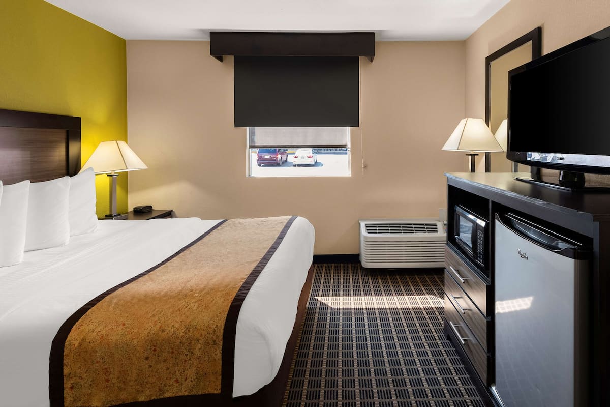 A hotel room with a bed, a television, a desk with a lamp, and a window with a view of parked cars.