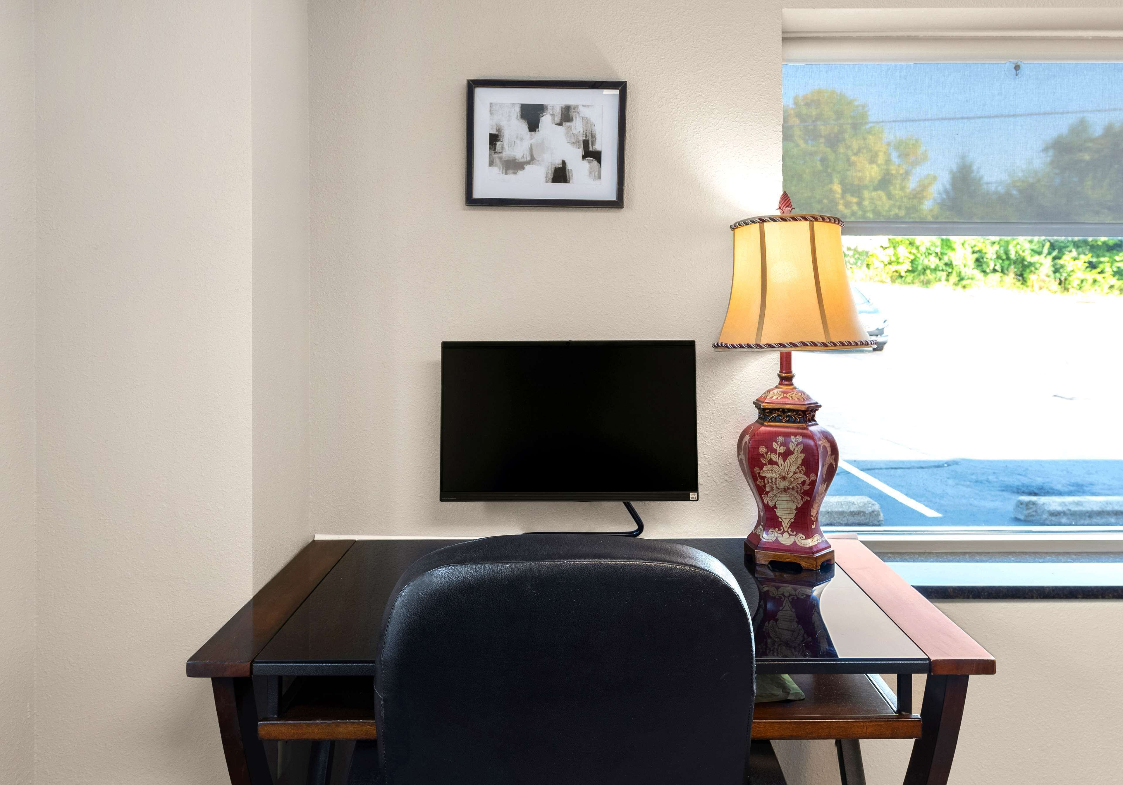 A room with a desk, a flat-screen TV, a lamp, and a window with a view of a parking lot.