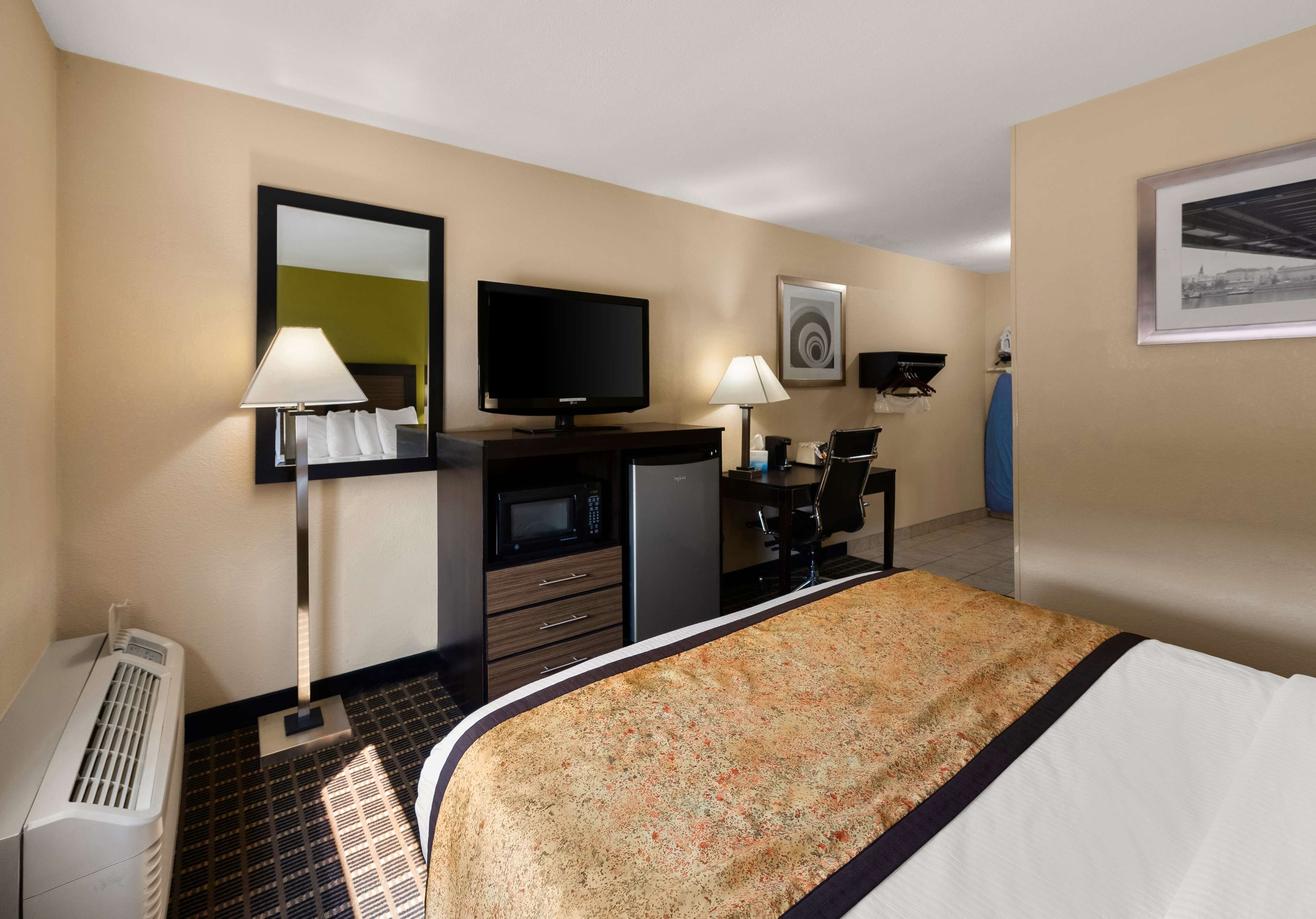Habitación de hotel con cama, televisor, escritorio con silla, espejo, lámpara y calefactor.