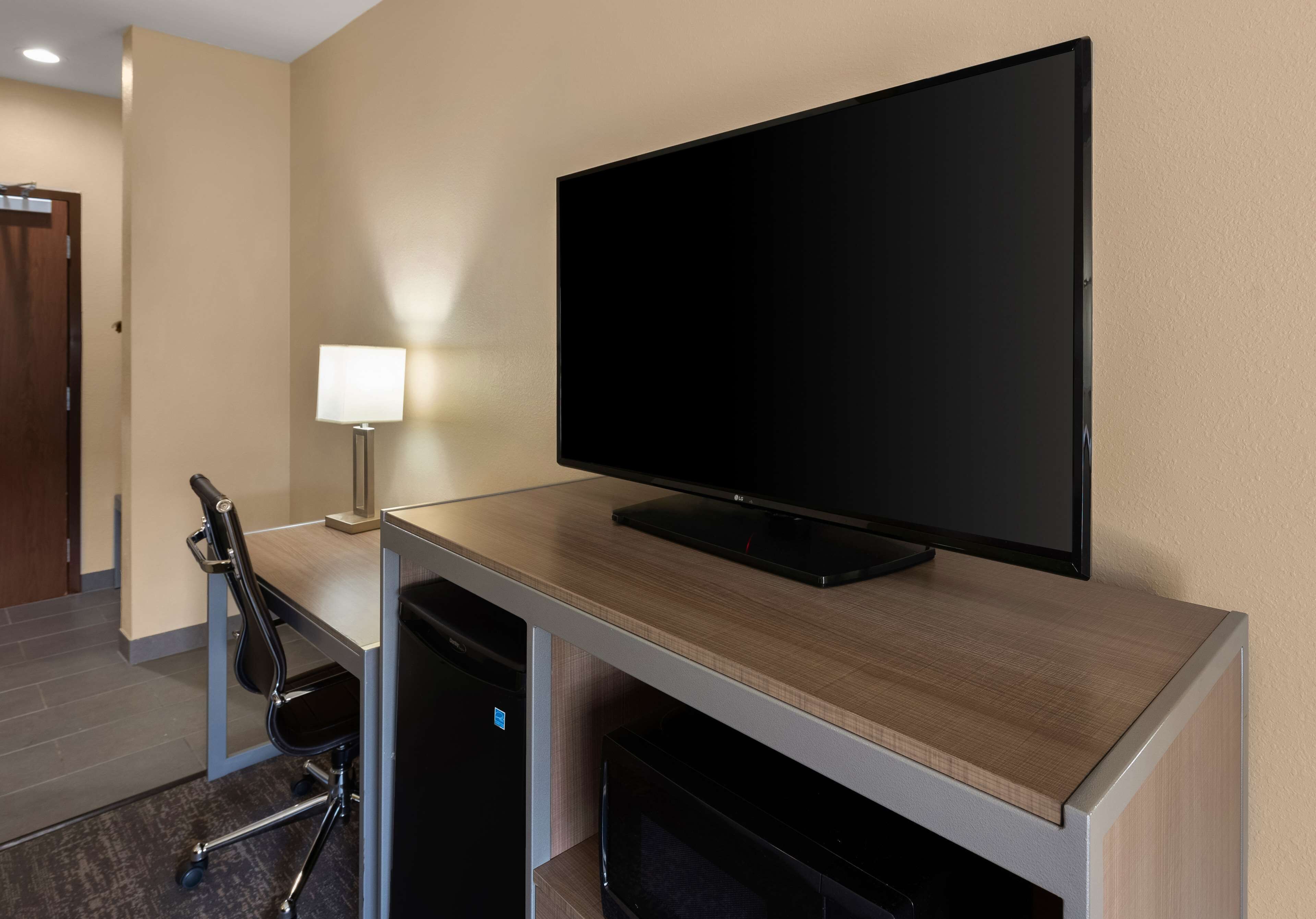 Habitación de hotel con televisor de pantalla plana montado en un escritorio de madera, una lámpara de escritorio, una silla de oficina negra y un microondas debajo del escritorio.