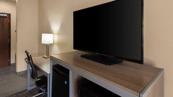 A hotel room with a flat-screen TV mounted on a wooden desk, a desk lamp, a black office chair, and a microwave below the desk.