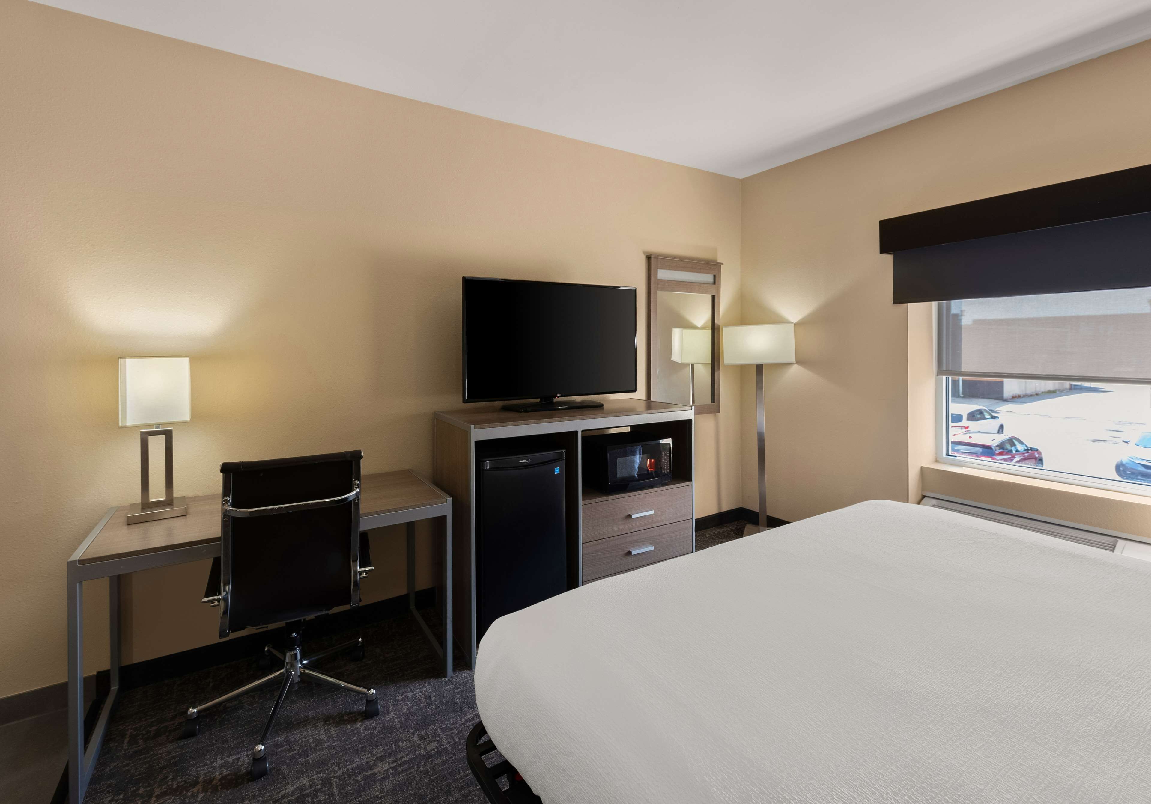 A hotel room with a bed, a desk with a chair, a television, a microwave, and a window with a view of a parking lot.