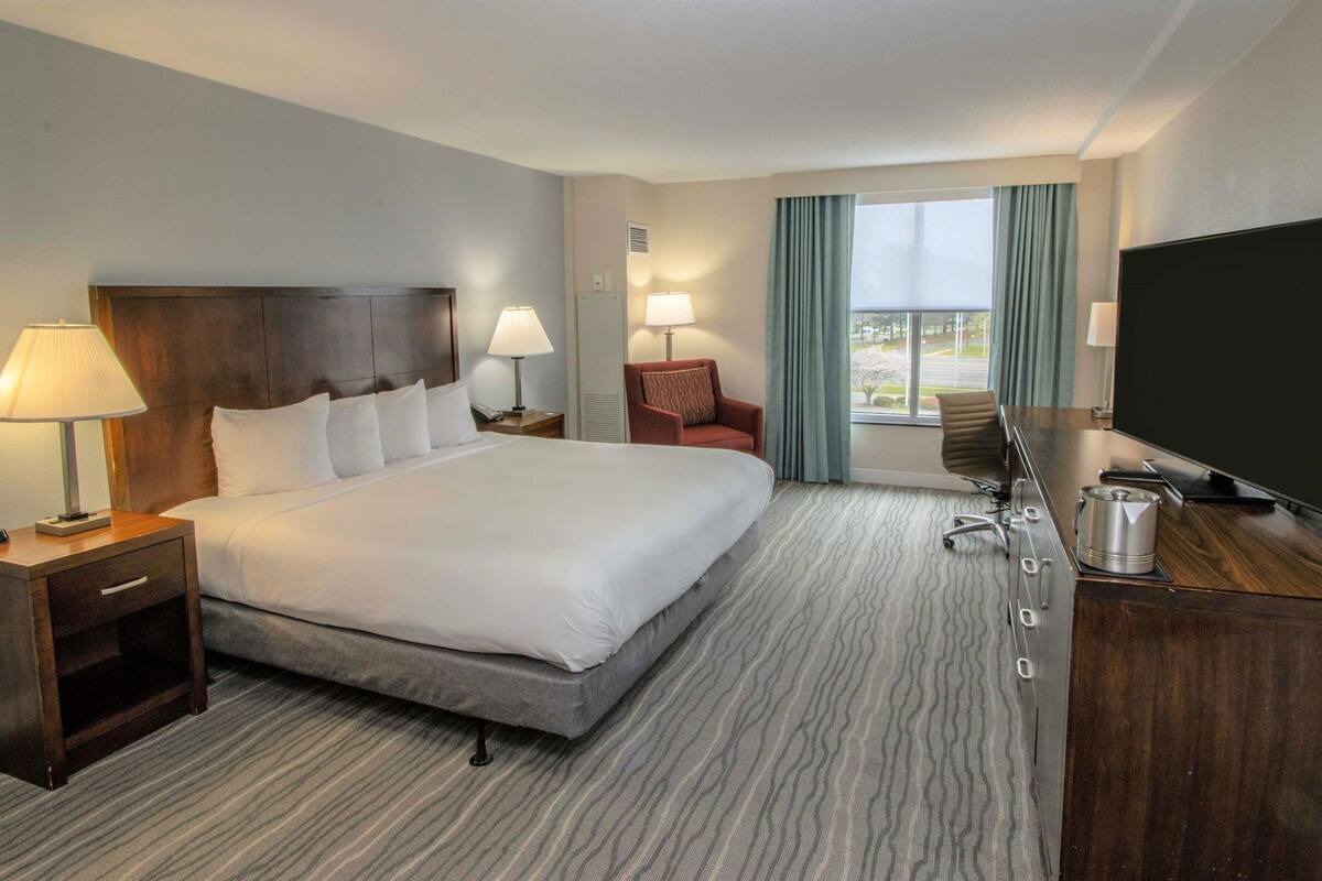 Quarto de hotel com uma cama grande, uma mesa com televisão de tela plana, uma cadeira, um abajur e uma janela com cortinas.