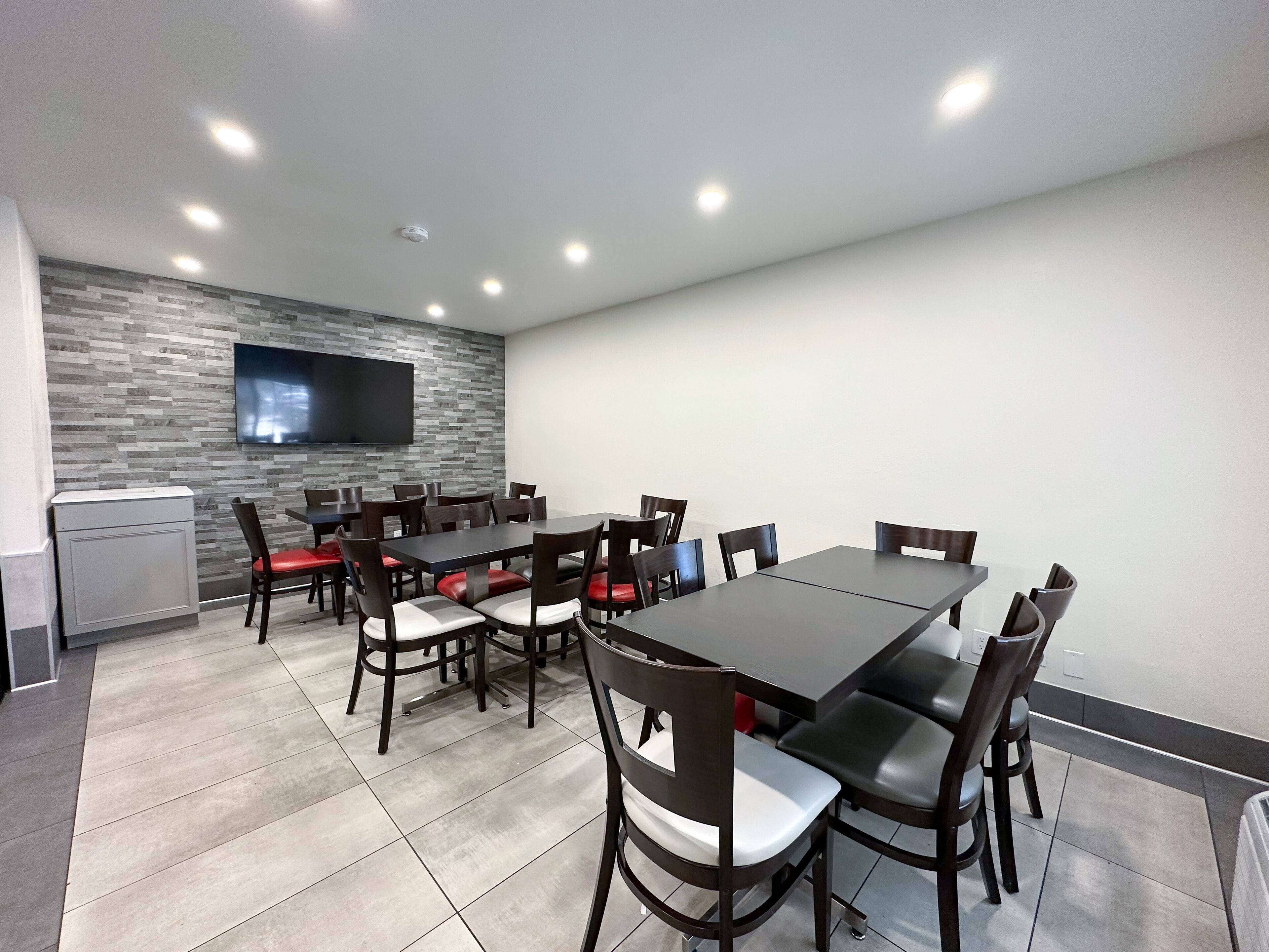 A modern dining area with a long table and chairs, a flat-screen TV mounted on a textured wall, and recessed ceiling lighting.