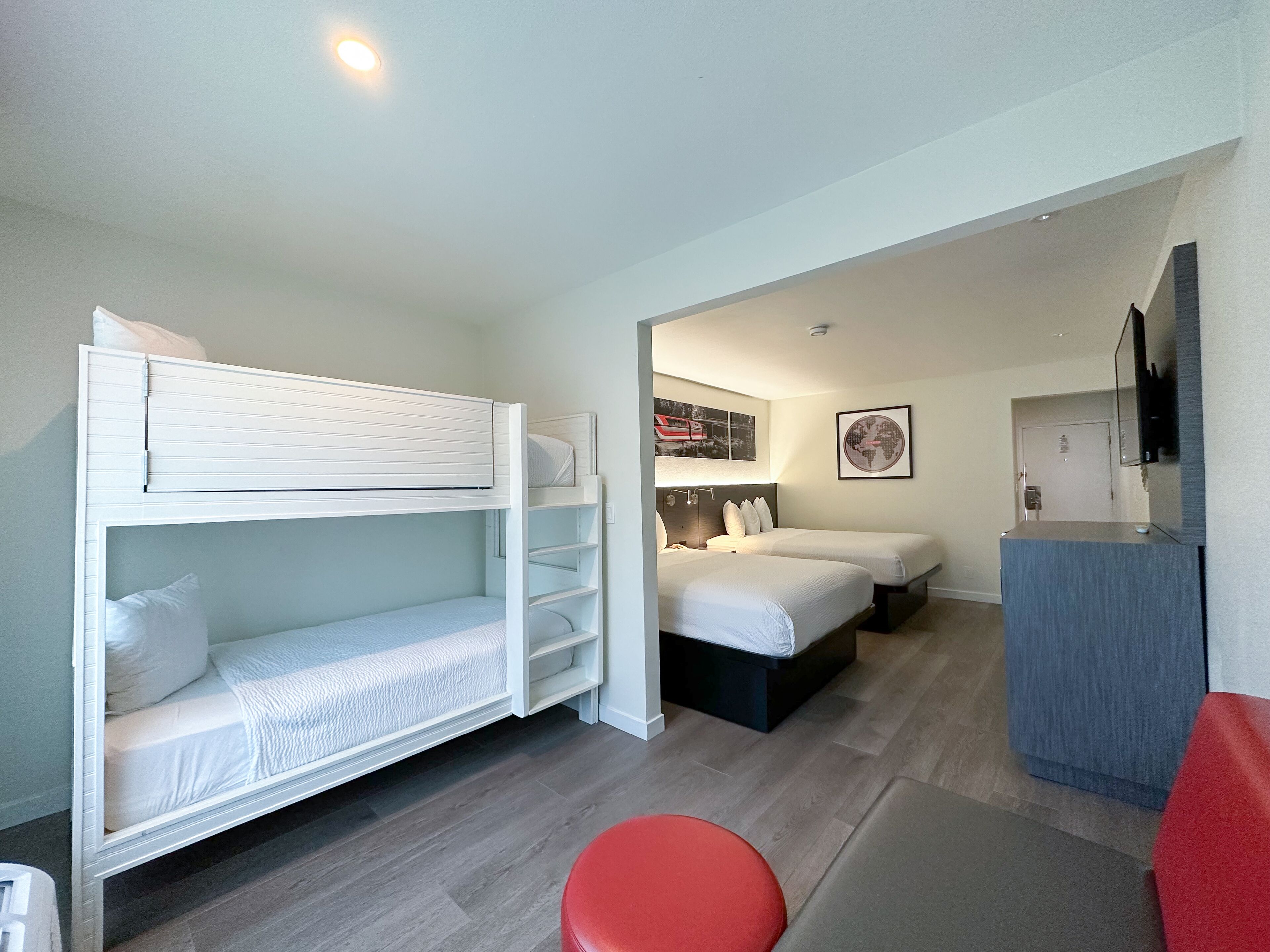 A hotel room with a bunk bed, two single beds, a desk, and a red stool.