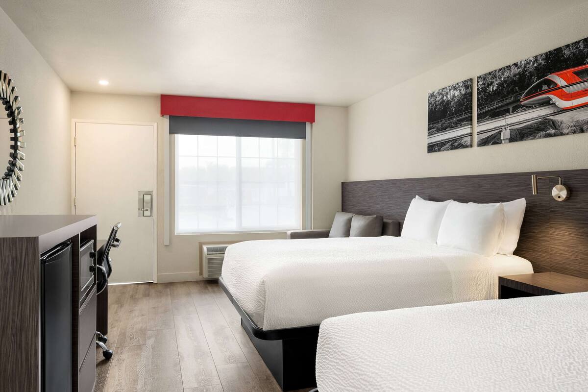 A hotel room with two beds, a desk, a chair, a window with a red valance, and a wall-mounted picture of a train.