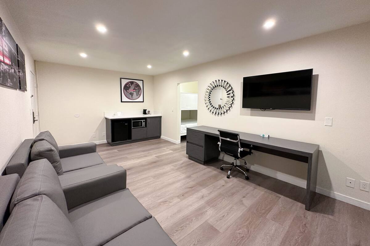 A modern living room with a gray sofa, a desk with a chair, a TV, and a kitchen area with a microwave.