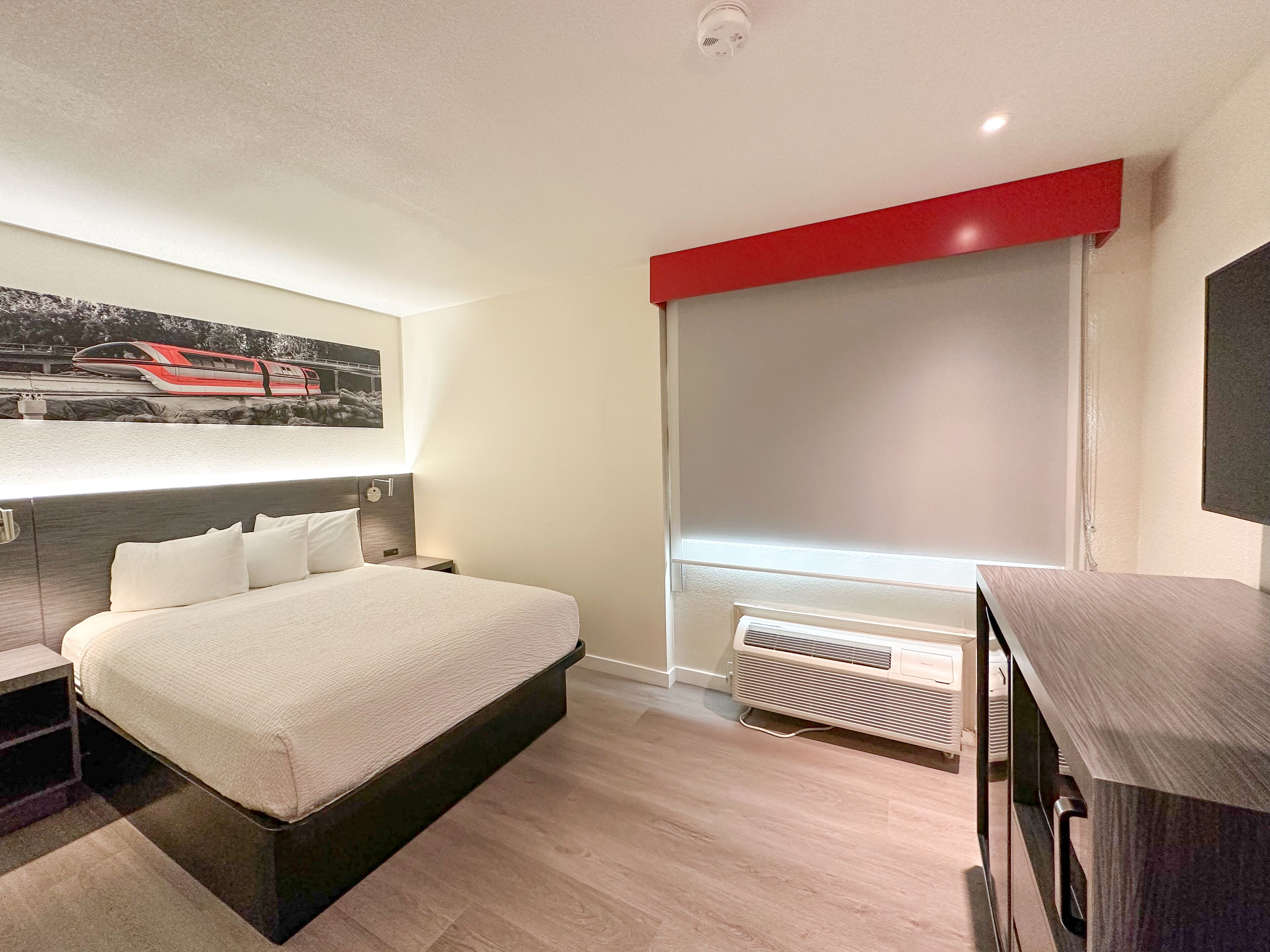 A modern bedroom with a large bed, a bedside table, a nightstand, a wall-mounted air conditioning unit, and a window with a red blind.