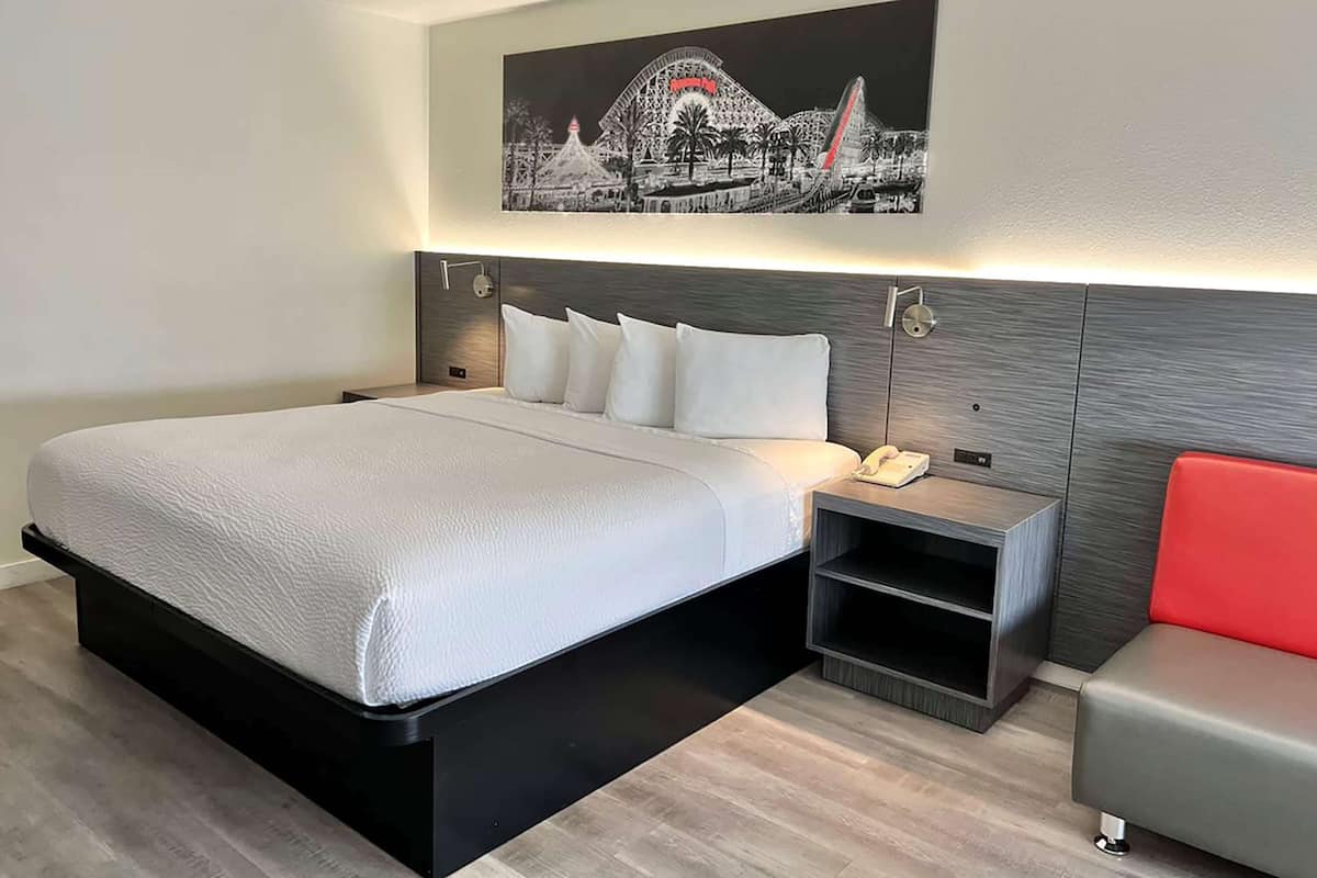 A modern hotel room with a large bed, a bedside table with a phone, a small shelf, and a red chair.