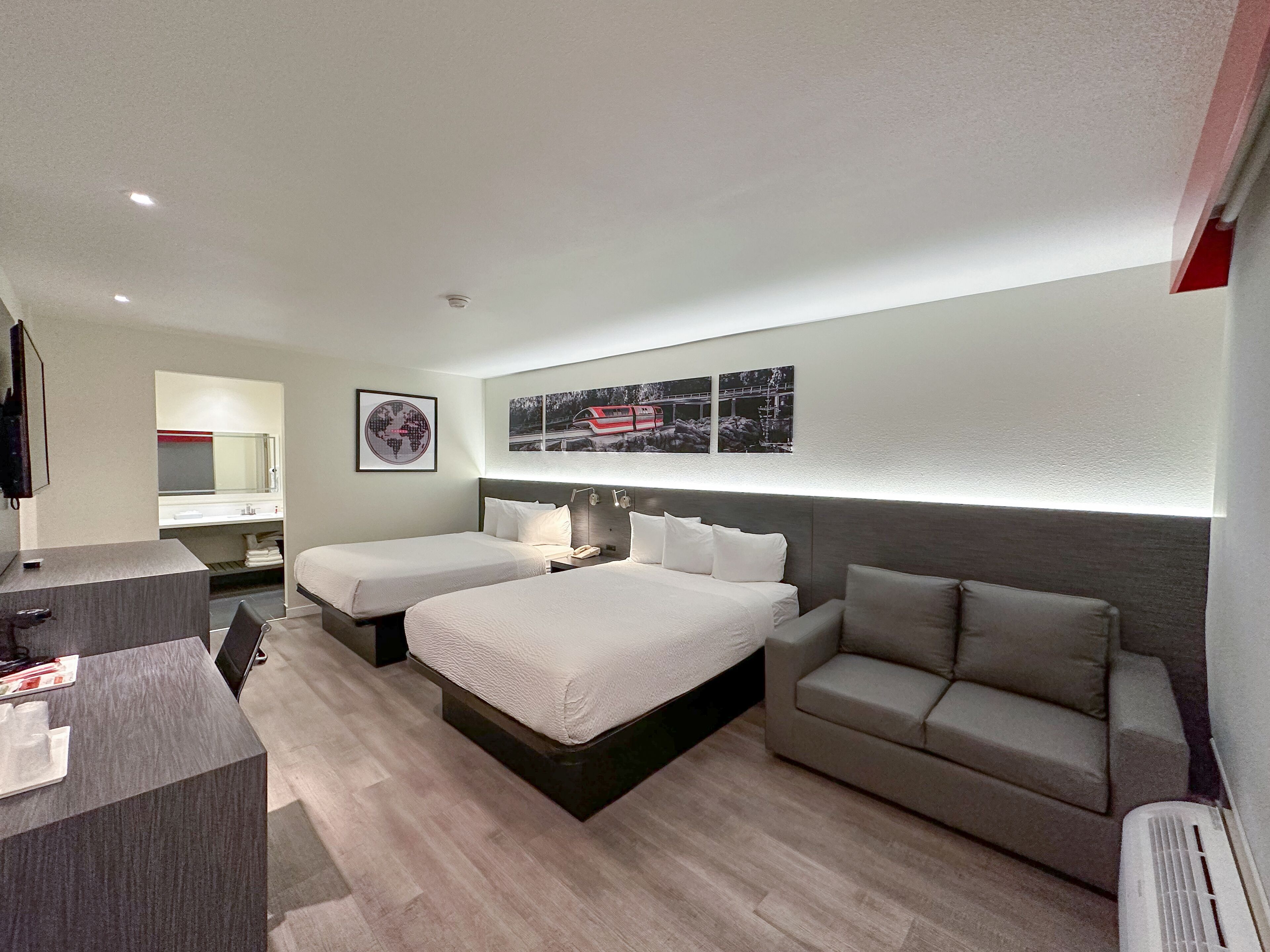 A modern hotel room with two beds, a sofa, a desk, and a bathroom.