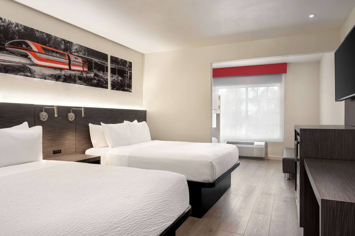 A hotel room with two beds, a large window, a flat-screen TV, and a painting of a train on the wall.