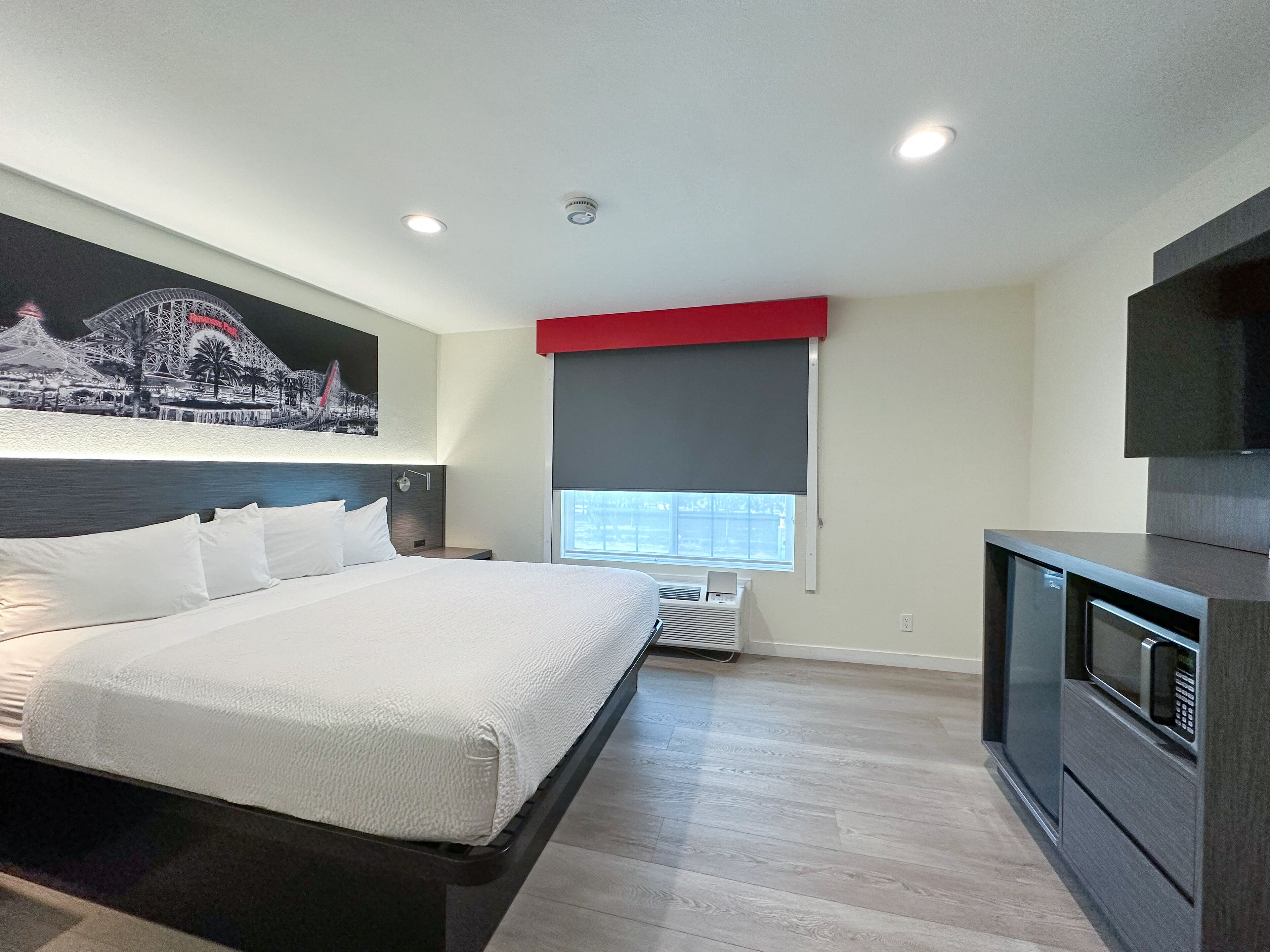 A modern hotel room with a large bed, a flat-screen TV, a window with a red blind, and a wall-mounted air conditioning unit.