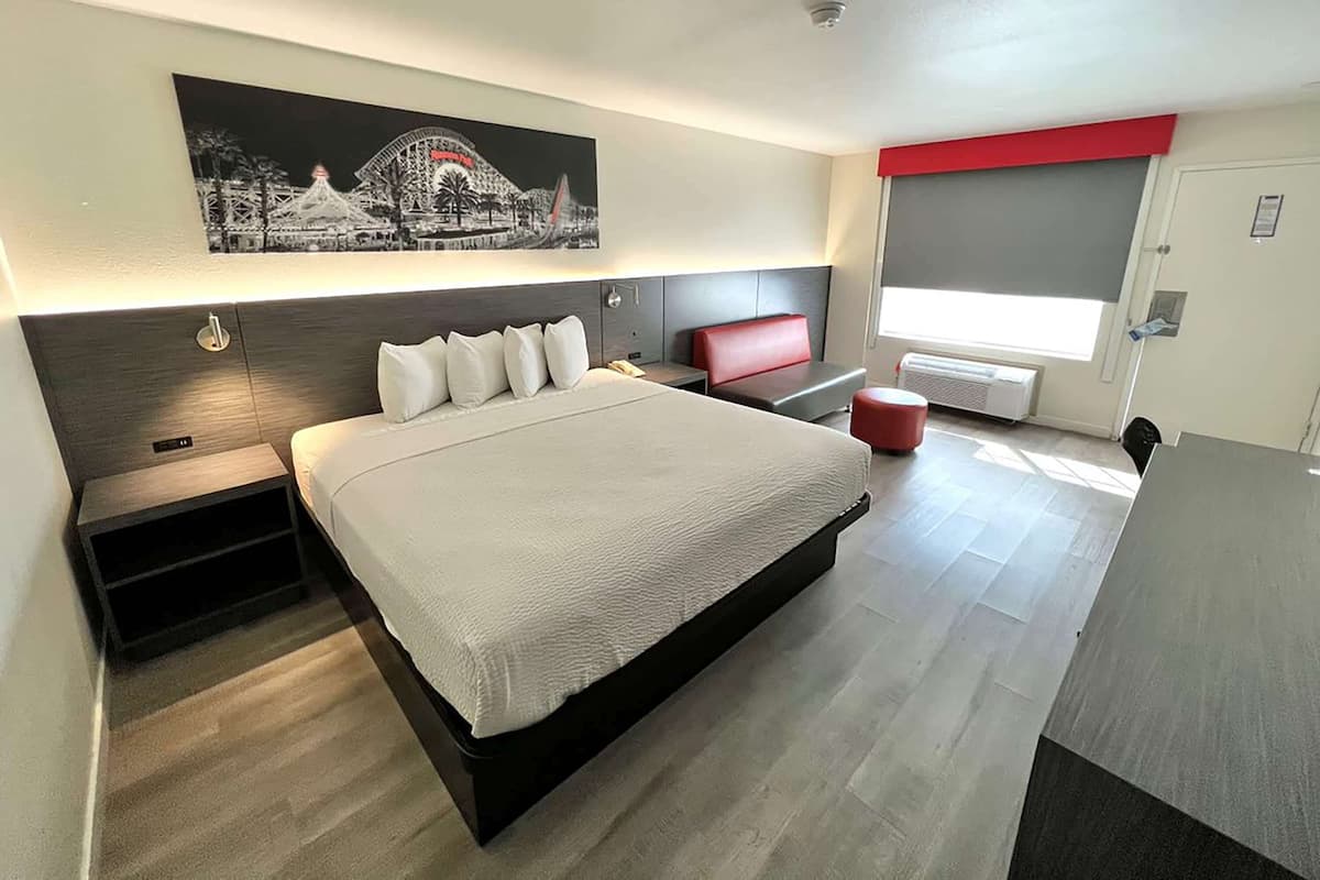 A modern hotel room with a large bed, a red bench, a desk, and a large window with a view of a building.