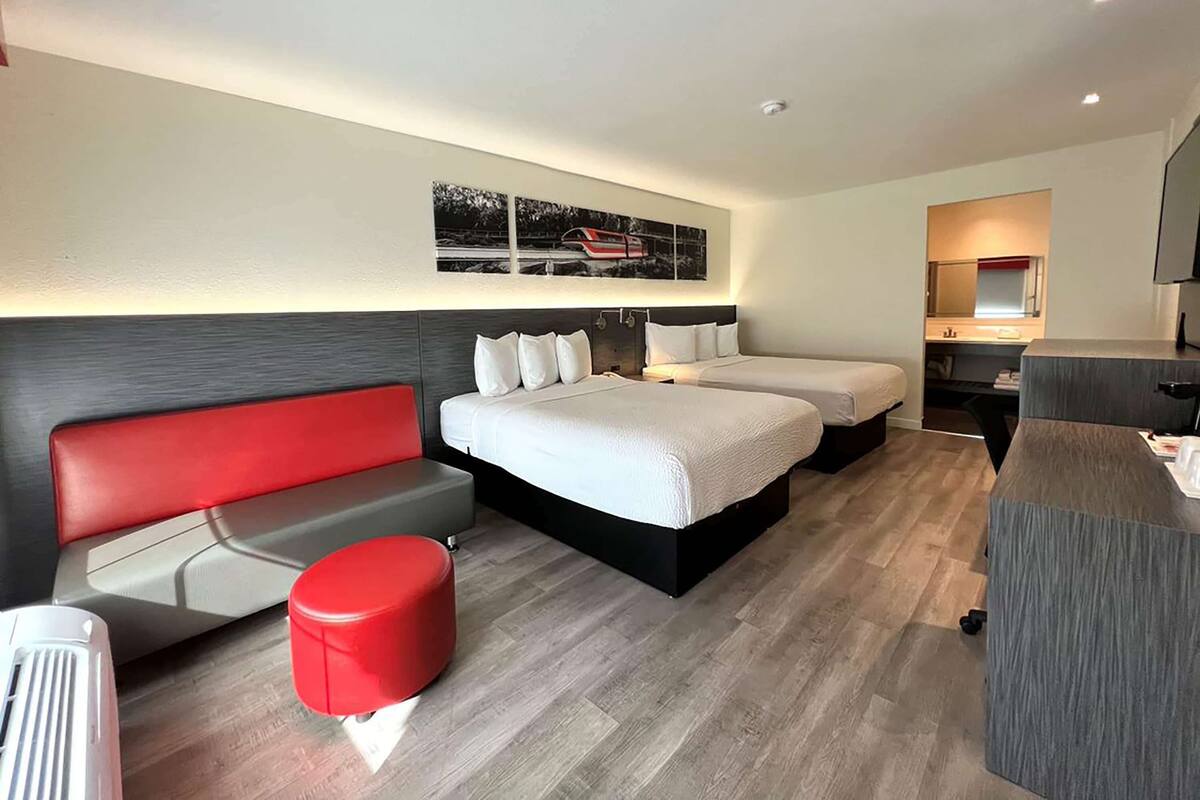 A modern hotel room with a large bed, a red sofa, a small red ottoman, and a desk with a mirror.
