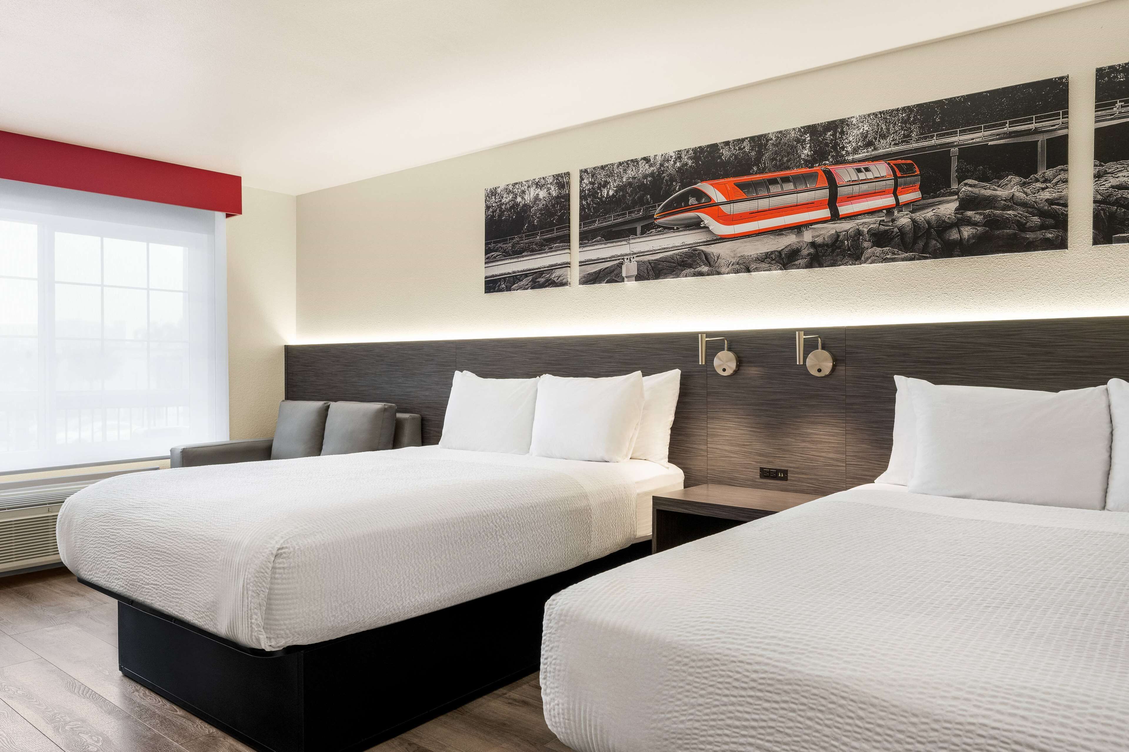 A hotel room with two beds, a large window, a modern headboard, and a wall-mounted artwork of a train.