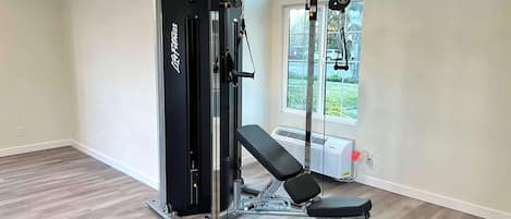 A gym with a weightlifting station, a bench, and a window with a view of greenery.