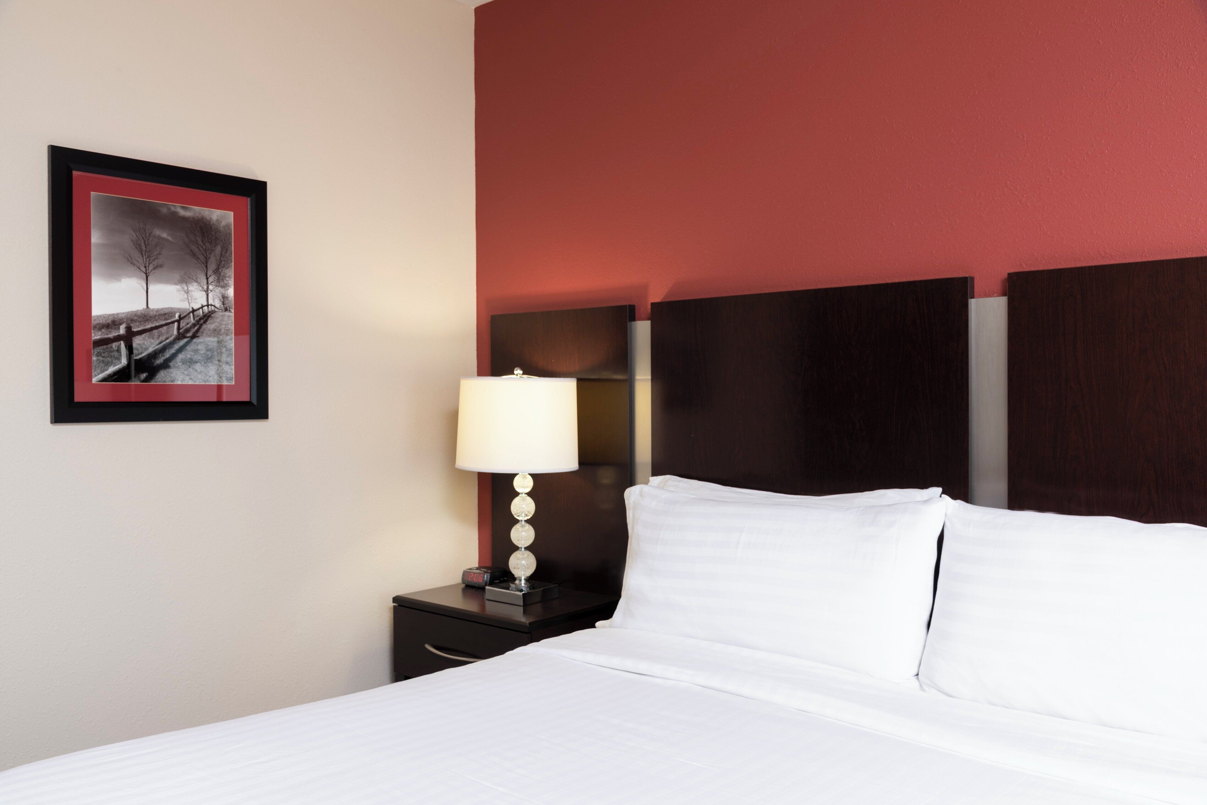 Habitación de hotel con cama, mesita de noche con lámpara, reloj y un cuadro en la pared.