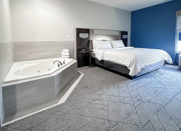 Quarto de hotel moderno com uma banheira grande, cama com roupa de cama branca e uma parede azul de destaque.