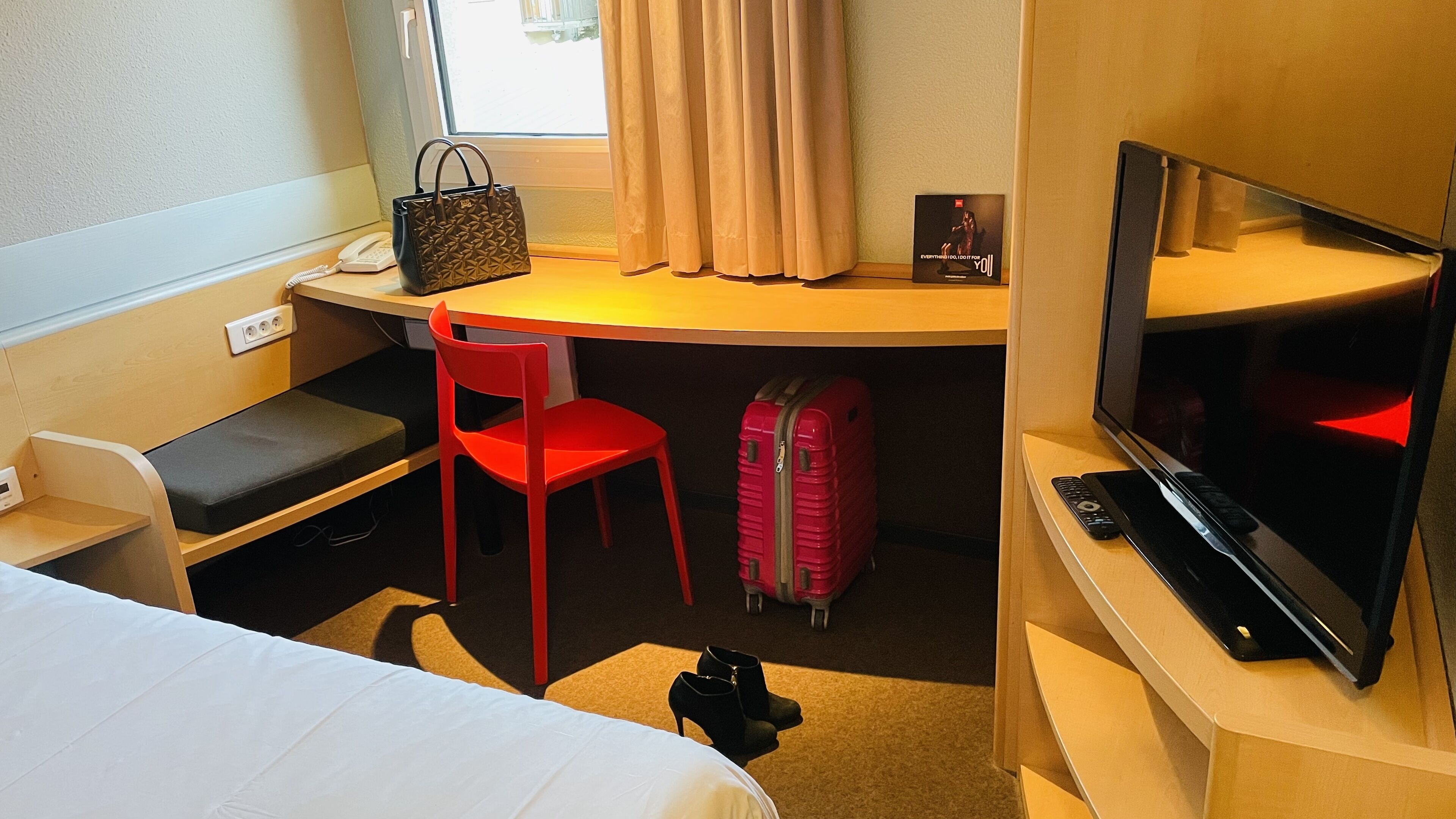 Une chambre d’hôtel avec un lit, un bureau et une chaise, une télévision, une valise et un sac à main.
