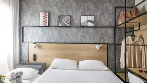 Une chambre d’hôtel moderne dotée d’un grand lit, d’une tête de lit en bois, d’un bureau avec une chaise, d’un porte-manteau et d’une étagère où sont exposées des œuvres d’art.