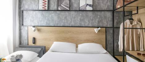 Una habitación de hotel moderna con una cama grande, un cabecero de madera, un escritorio con una silla, un perchero y una estantería con obras de arte.