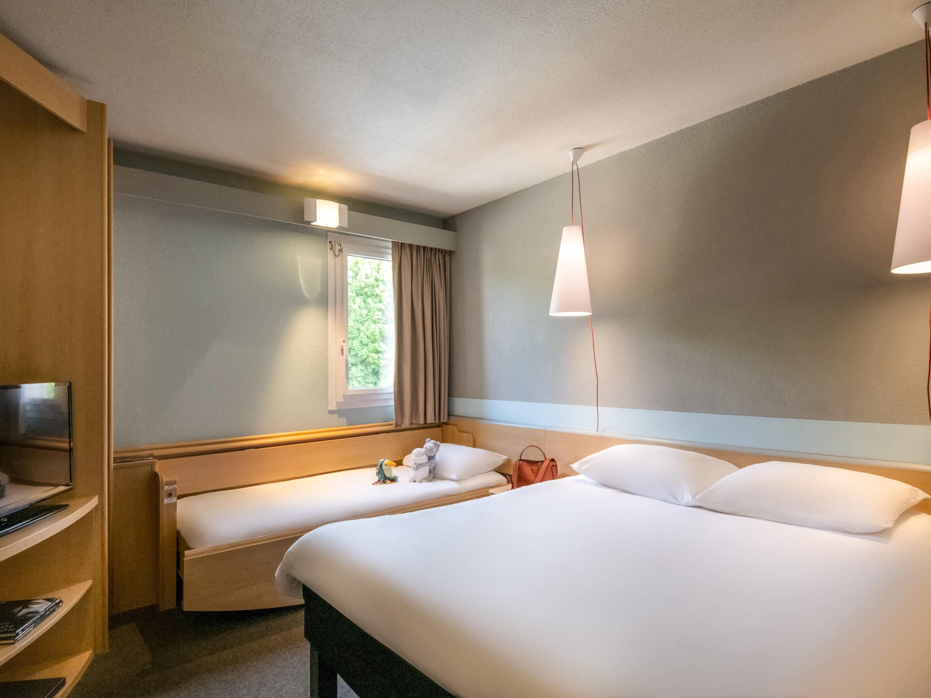 Habitación de hotel con cama, mesita de noche, televisión, ventana con cortinas y lámpara colgante.