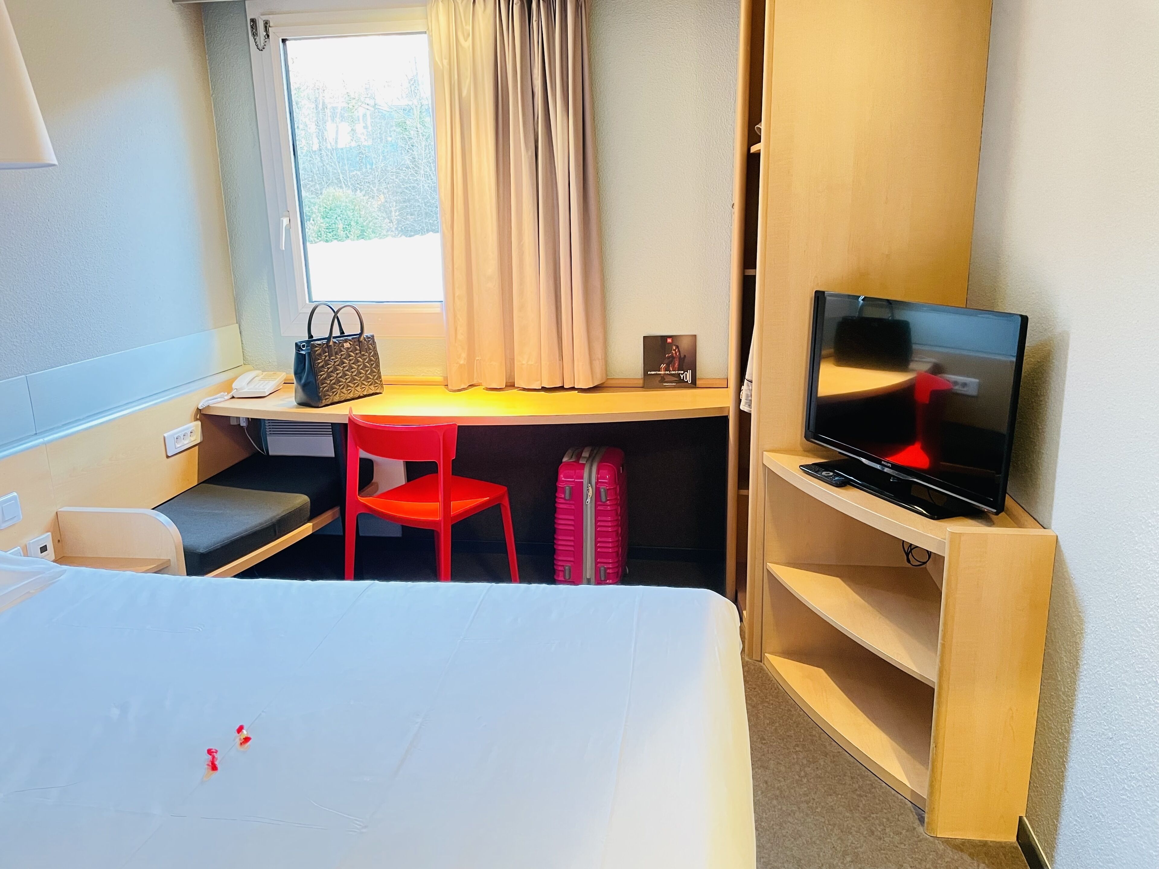 A hotel room with a bed, a desk with a chair, a TV, a suitcase, and a window with curtains.