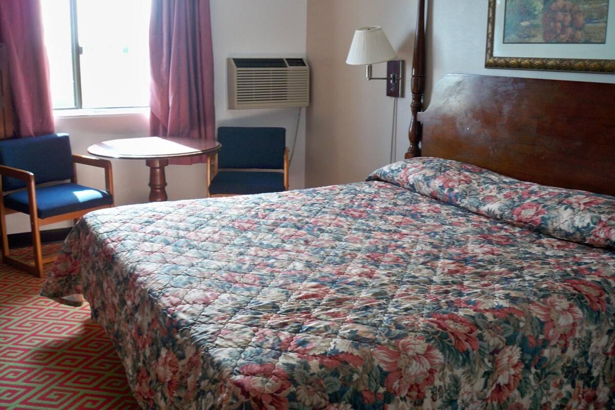Une chambre d’hôtel avec une couvre-lit à motifs floraux, une table ronde avec deux chaises, un climatiseur, une lampe et un tableau au mur.