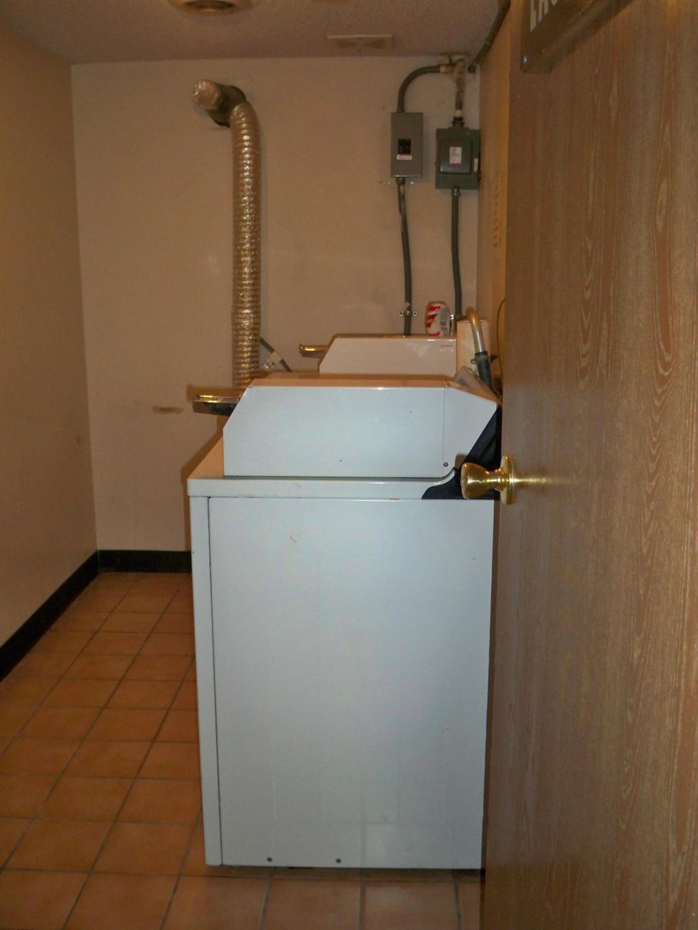 Ett litet, vitt förråd med en varmvattenberedare, en ventilationsslang och en dörr med ett handtag i mässing.
