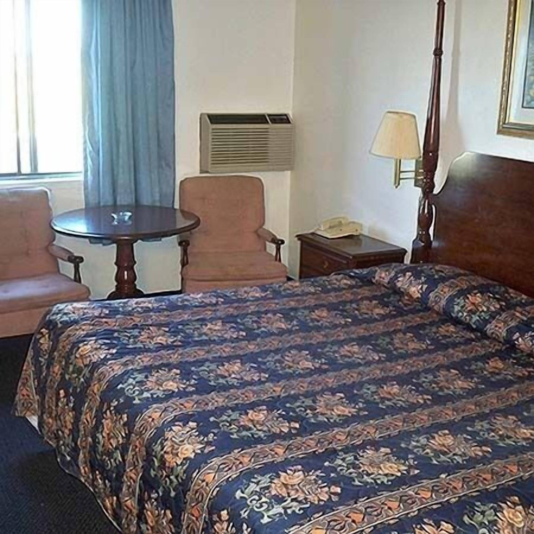 A hotel room with a bed, a chair, a small table, a lamp, and an air conditioning unit.