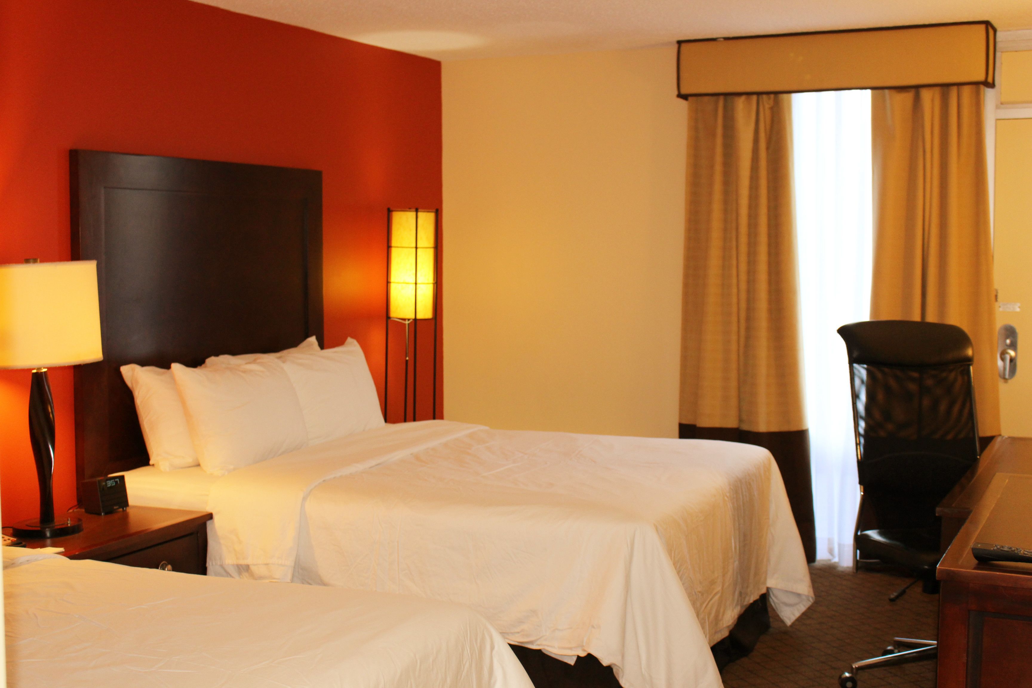 A hotel room with two beds, a desk, a chair, and a lamp.