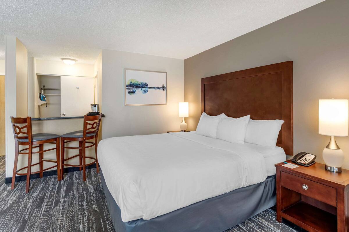 A hotel room with a large bed, a desk with two chairs, a nightstand with a lamp, and a painting on the wall.
