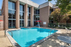 A swimming pool with clear blue water, surrounded by a brick building with large windows and a covered seating area with umbrellas.