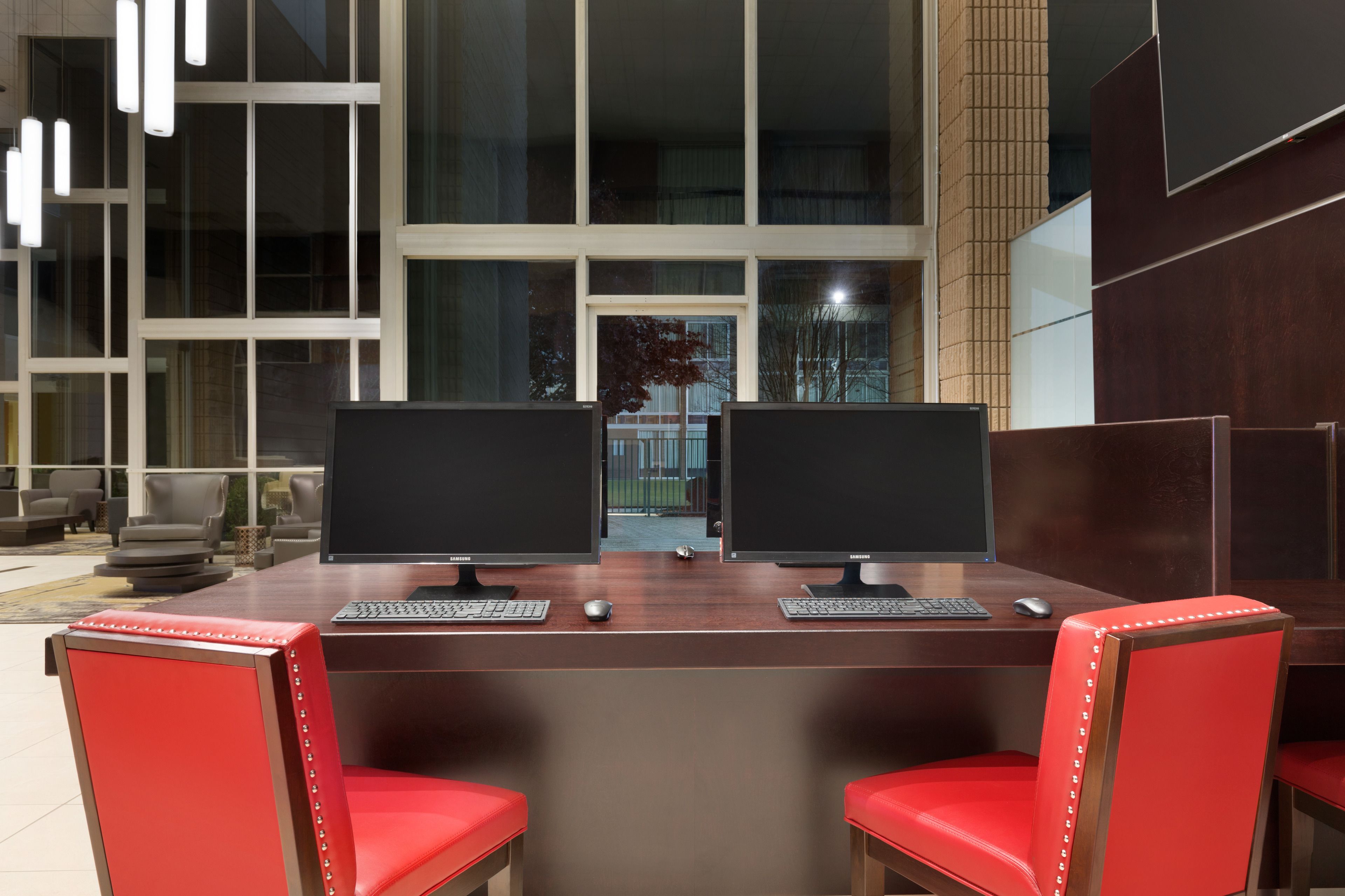 A modern office space with two desks, each with a computer monitor, keyboard, and mouse. There are red chairs with metal studs. Large windows provide a view of the outside.