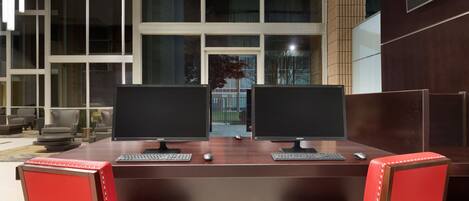 A modern office space with two desks, each with a computer monitor, keyboard, and mouse. There are red chairs with metal studs. Large windows provide a view of the outside.