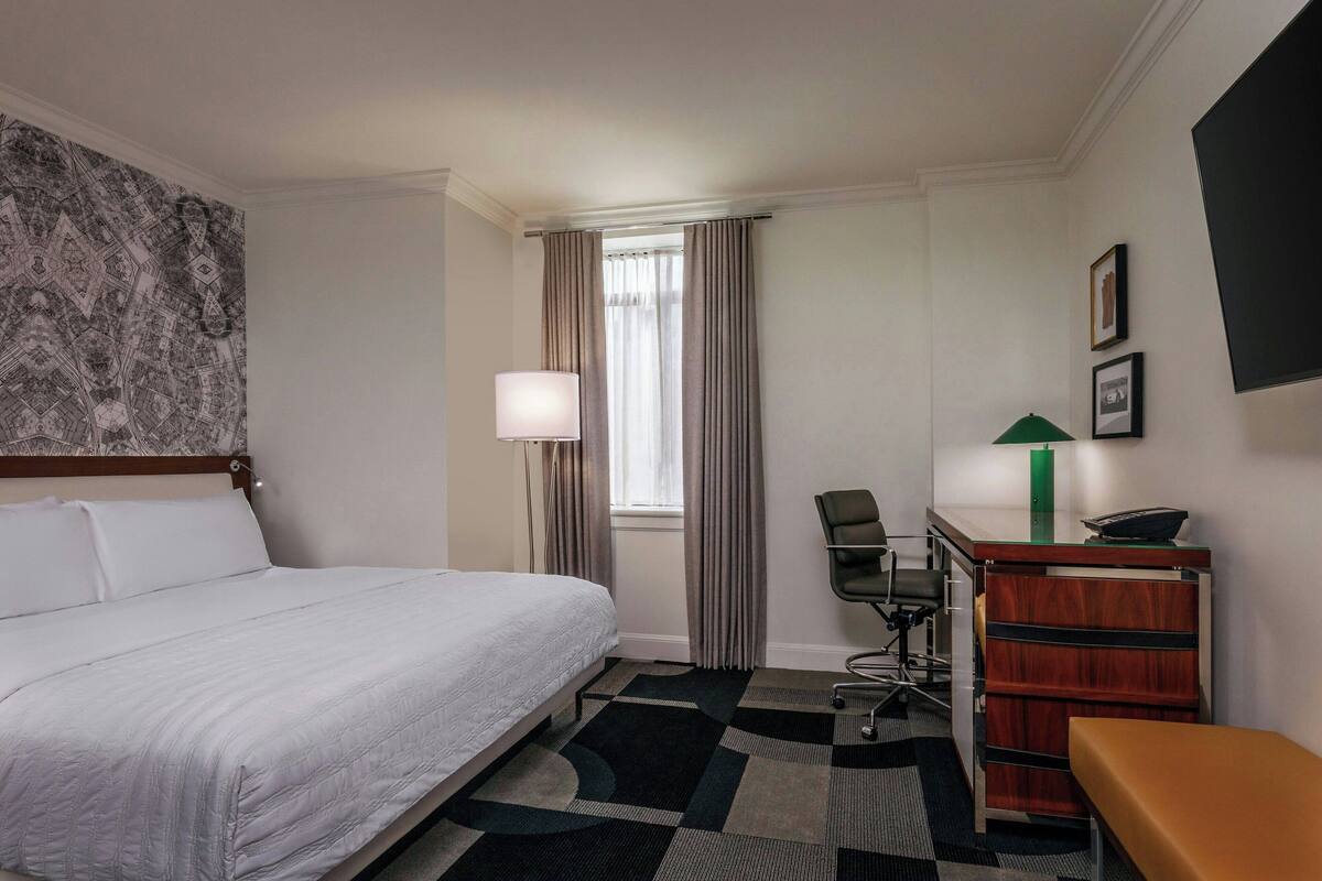 A hotel room with a bed, desk, chair, lamp, and a patterned wall.