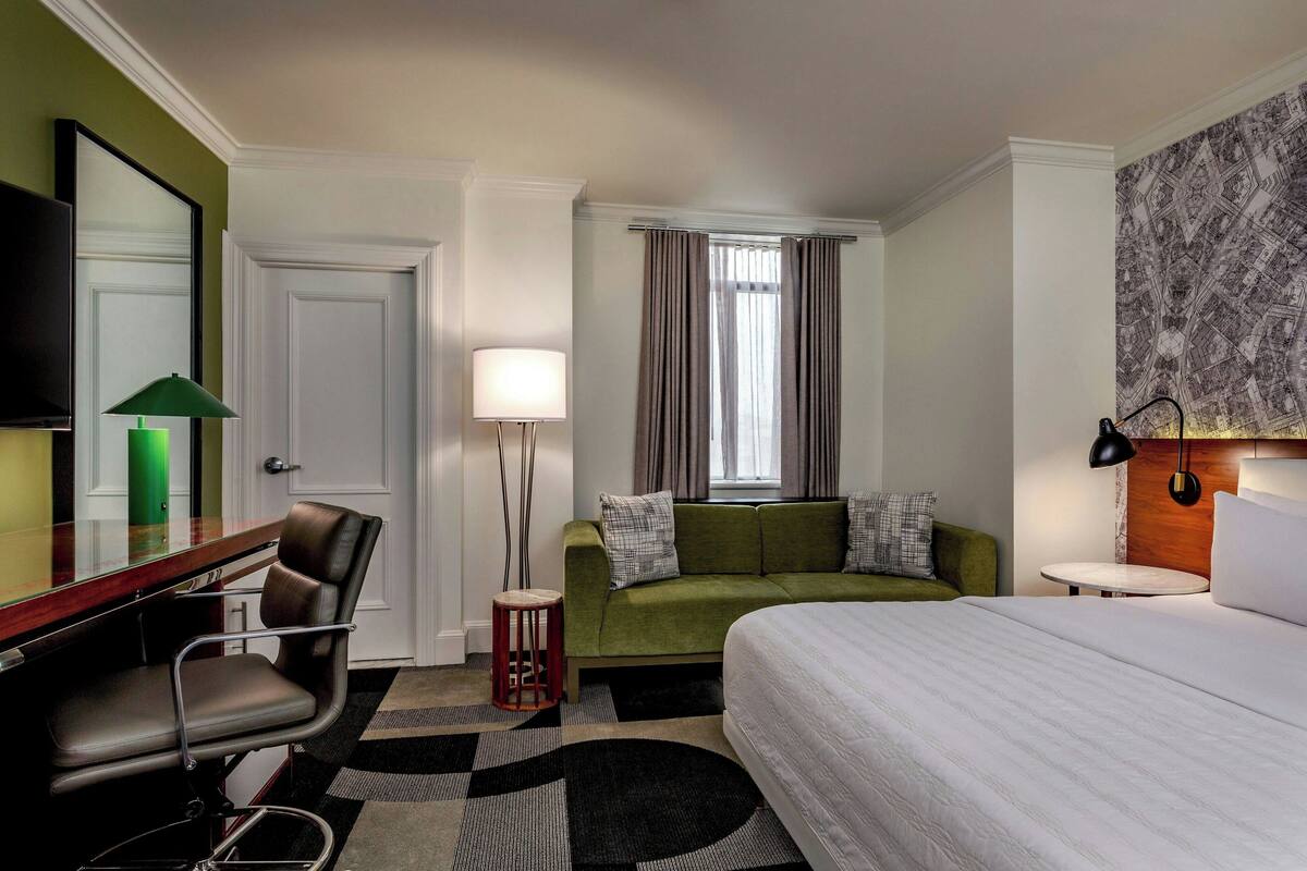 A hotel room with a bed, a desk with a chair, a sofa, a small round table, a lamp, and a large mirror.