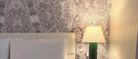 A neatly made bed with white linens, a green lamp on a bedside table, and a patterned wallpaper.