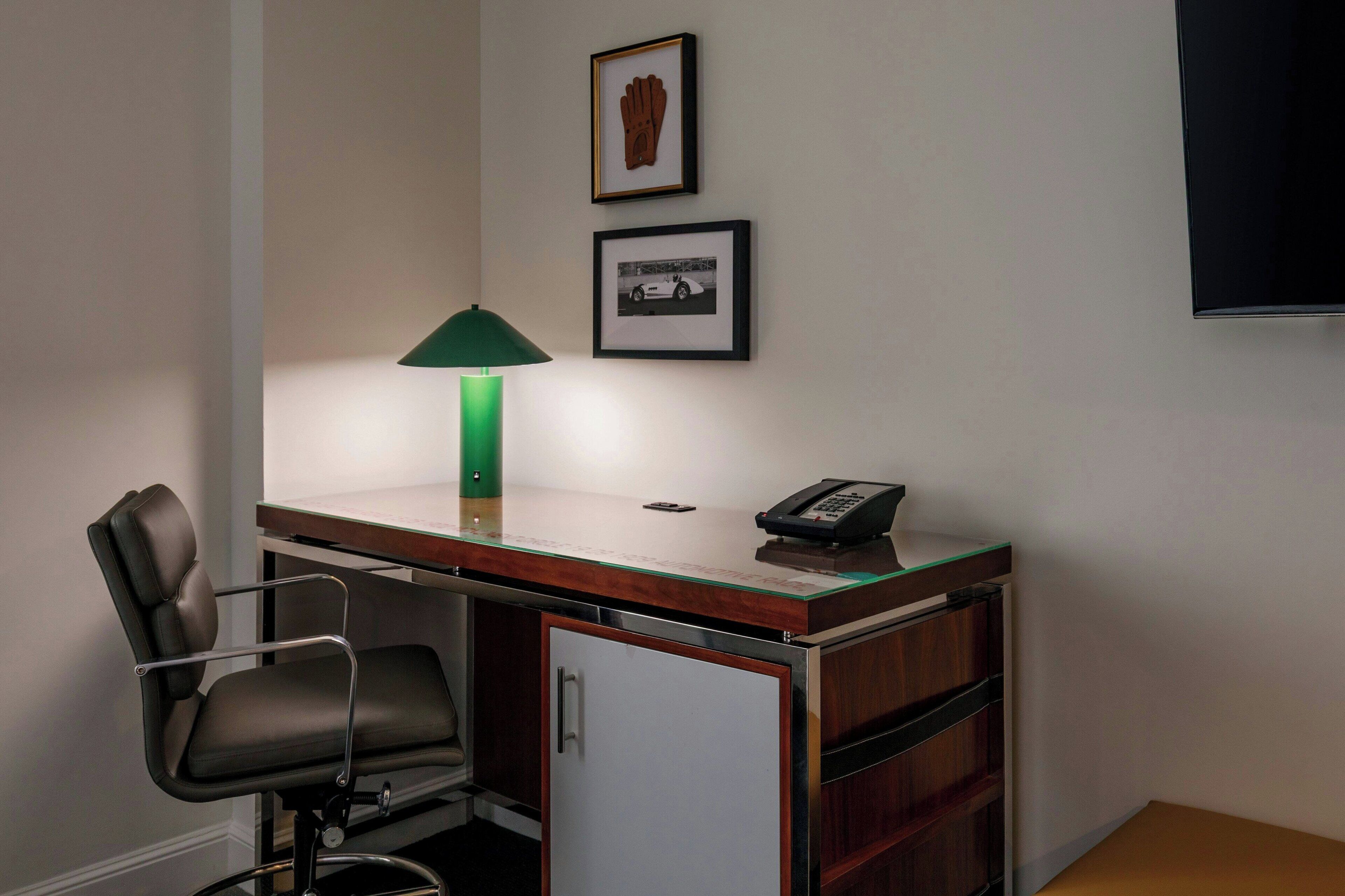 A small office space with a desk, chair, lamp, phone, and framed pictures on the wall.