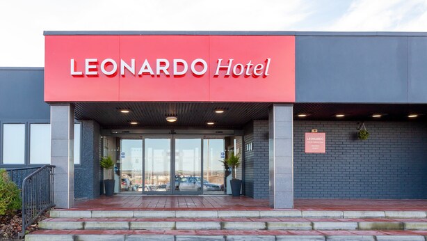 L’extérieur de l’hôtel LEONARDO se distingue par sa façade rouge, son entrée en verre et une enseigne indiquant qu’il s’agit d’un hôtel et d’un centre de conférences.