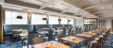A hotel dining area with multiple tables and booths, a patterned carpet, and a ceiling with recessed lighting.