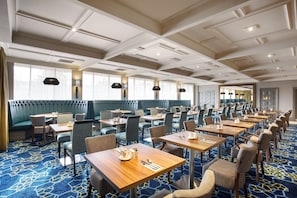 A hotel dining area with multiple tables and booths, a patterned carpet, and a ceiling with recessed lighting.