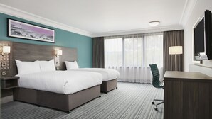 Une chambre d’hôtel avec un grand lit, un bureau et une chaise. Il y a une fenêtre avec des rideaux, un tableau au mur et une lampe sur le bureau.