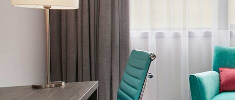 A hotel room with a desk, a teal chair, a lamp, a window with sheer curtains, and a striped carpet.