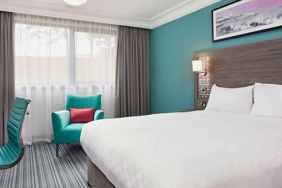 A hotel room with a large bed, a teal armchair, a window with sheer curtains, and a framed picture on the wall.