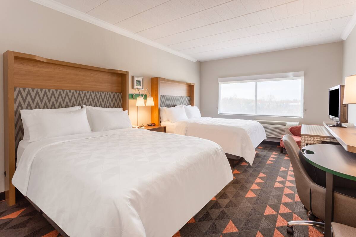 Ein Hotelzimmer mit zwei Betten, einem Schreibtisch, einem Stuhl, einer Lampe und einem großen Fenster.