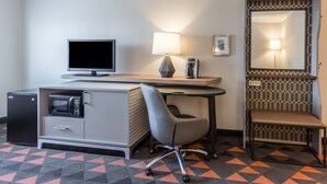Habitación de hotel con escritorio, una silla, refrigerador pequeño, microondas, lámpara, espejo y alfombra estampada.